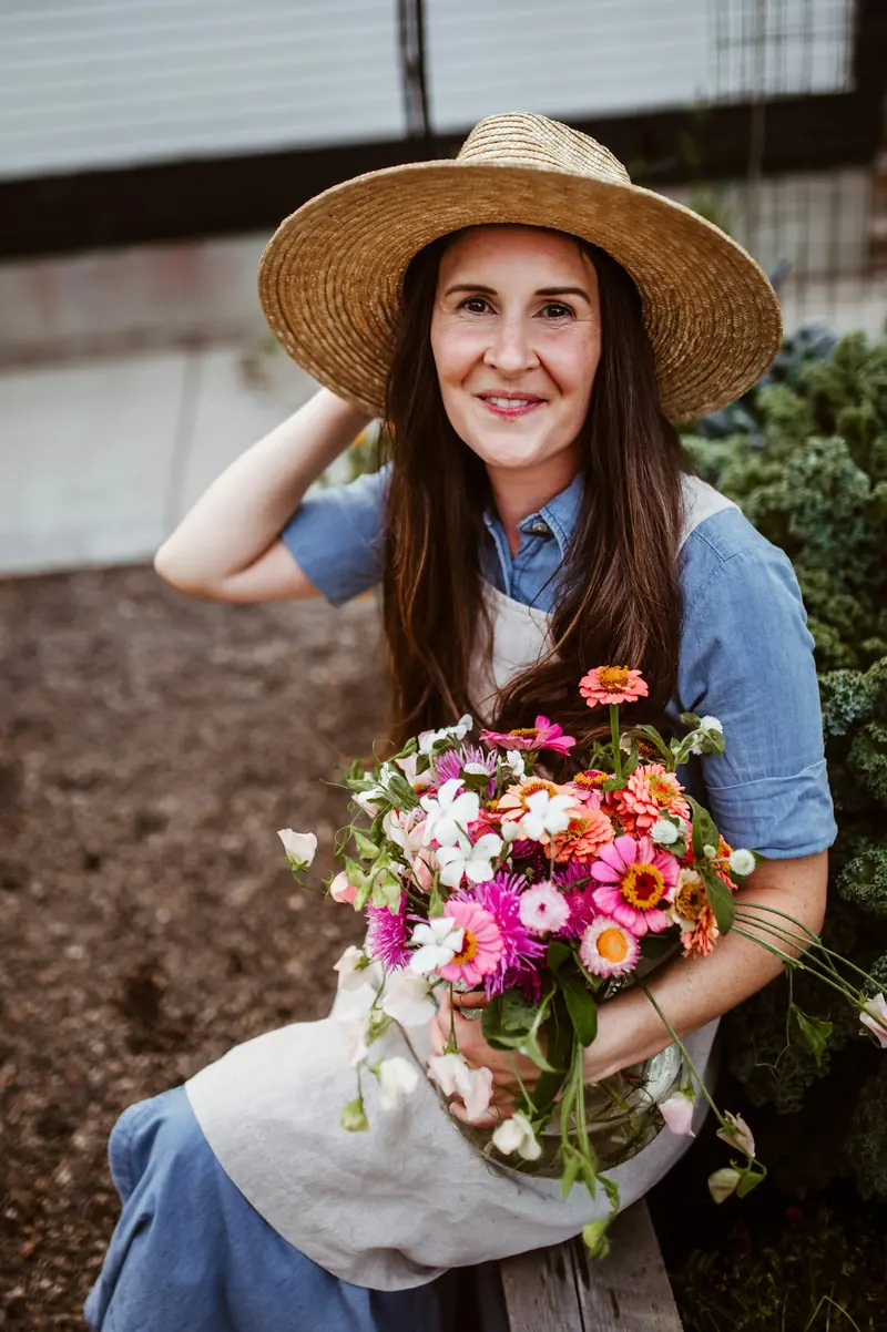 A flower farmer in a straw hat holding an armful of zinnias