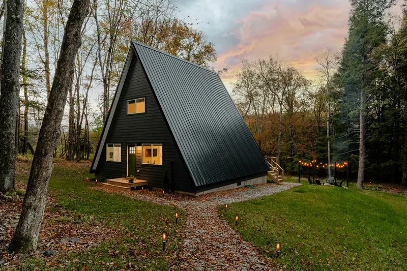 A small black A-frame cabin with lit windows in the forest at dusk
