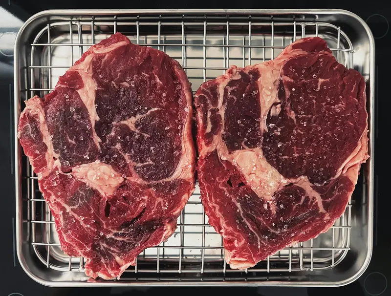 Two raw ribeye steaks salted on a rack