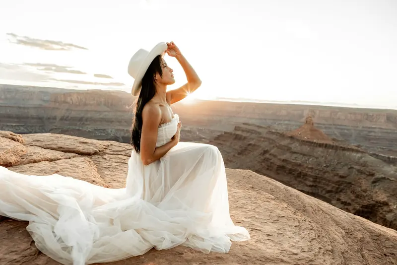A bride in a white dress and hat sitting on a rock at sunset