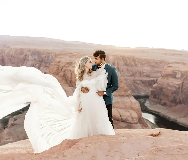 A bride and groom on a canyon rim with her dress blowing