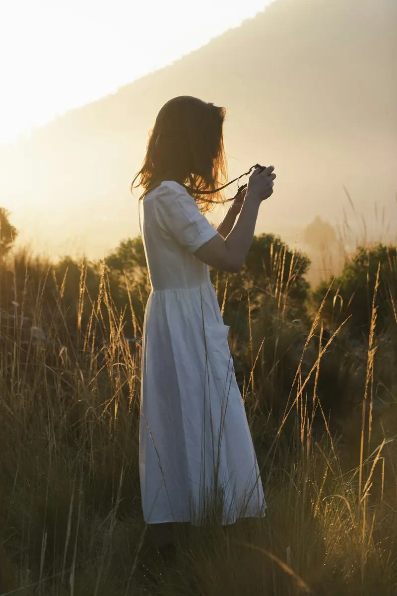 A woman in a white dress holding a camera in a field at sunset