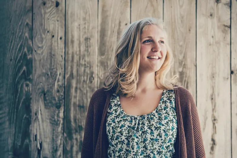 A smiling woman in a brown cardigan in front of a weathered wood wall