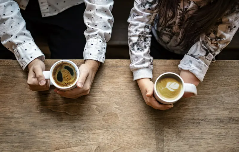 Two people holding cups of coffee across a wooden table